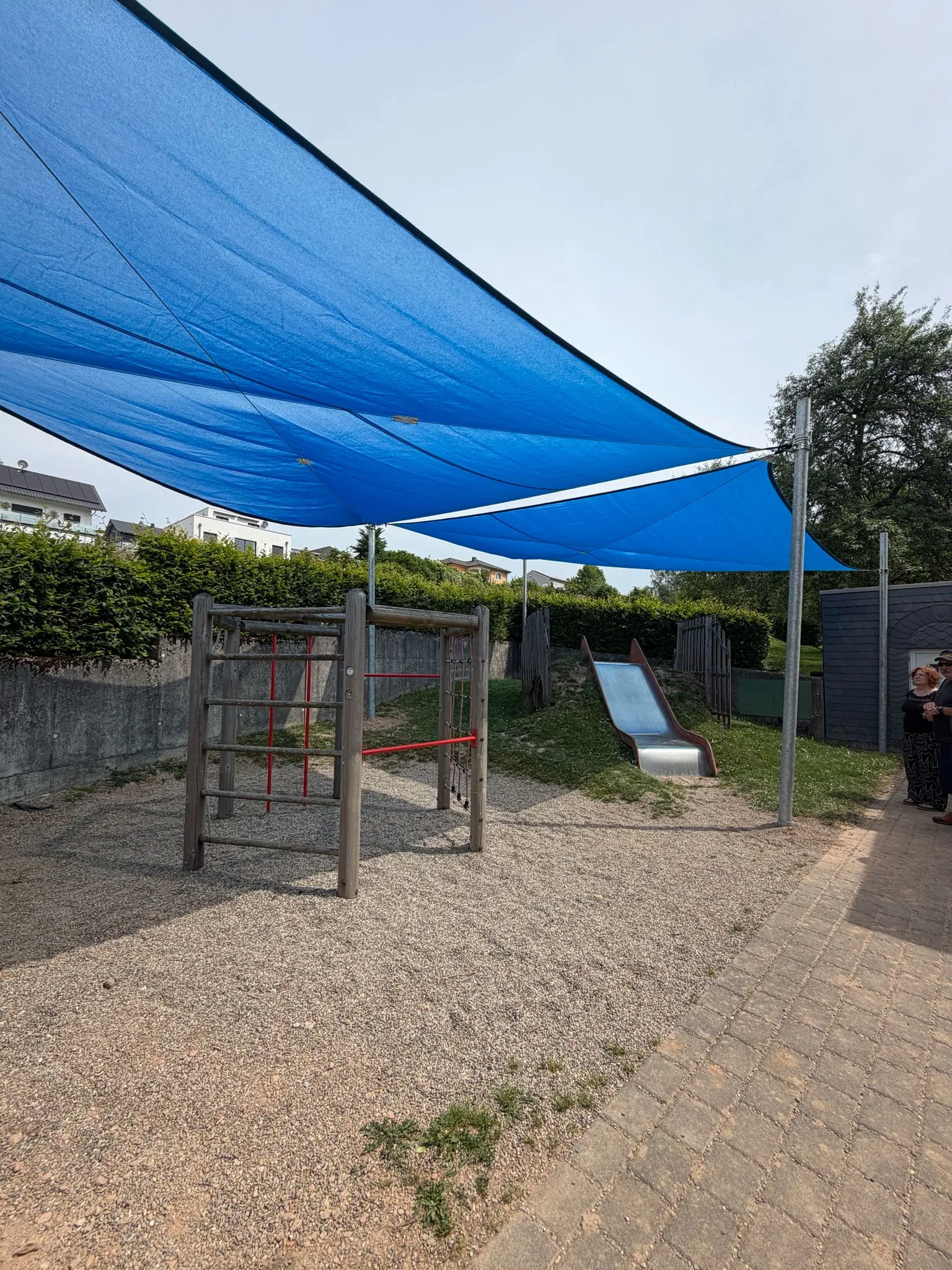Mehrere blaue Sonnensegel beschatten eine Holz-Kletteranlage mit Edelstahlrutsche, montiert von Moshrif Spielplatzbau